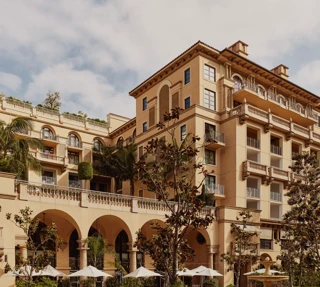 Arched terrace of The Maybourne Beverly Hills facing Beverly Canon Gardens, with palms and white umbrellas below.