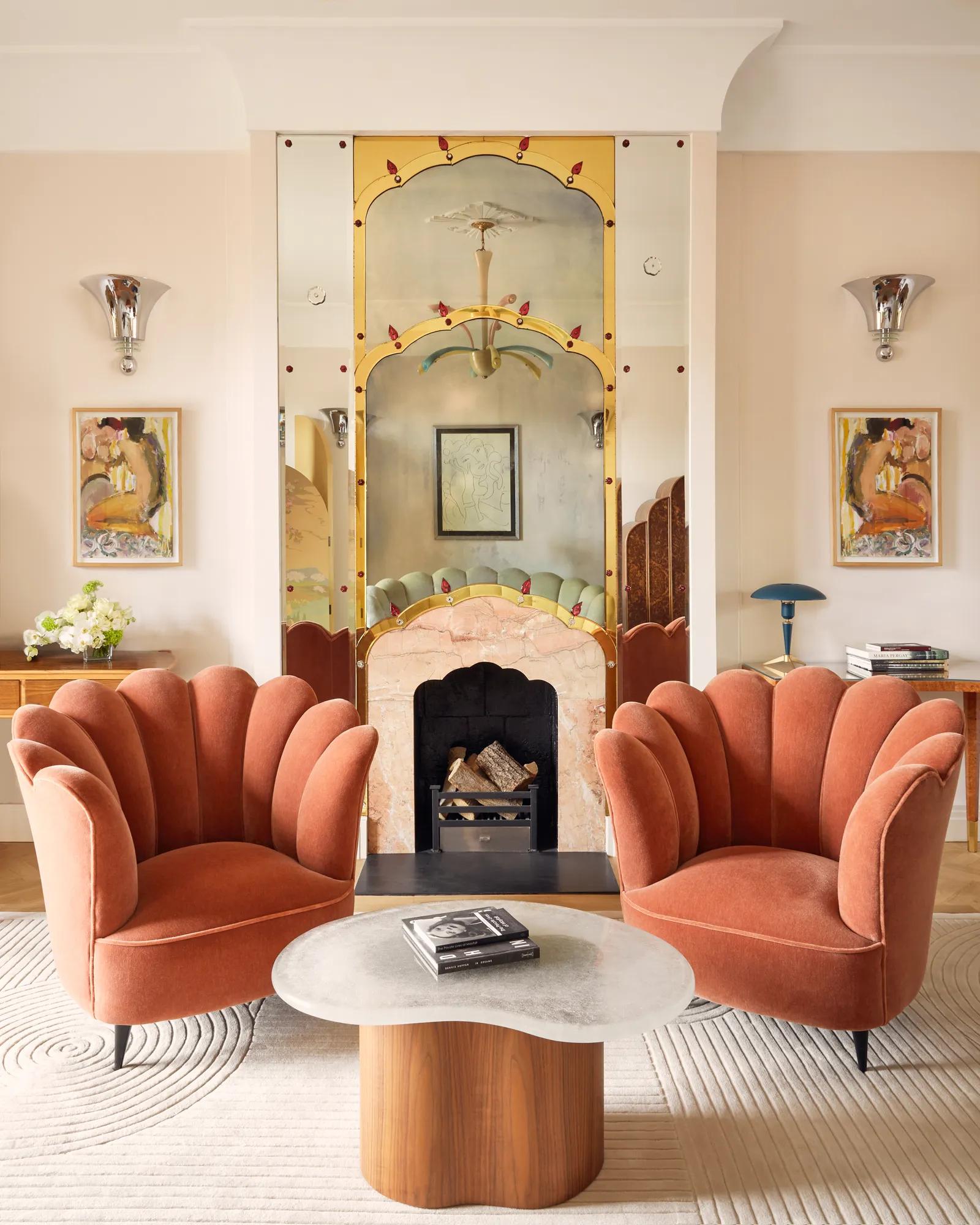 Art Deco-inspired sitting area with two coral velvet scallop armchairs and a marble coffee table, set before a decorative mirrored fireplace framed with painted detailing and flanked by colourful artwork.