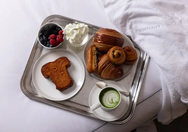 Breakfast tray on a bed with pastries, brioche toast, berries, a cup of green matcha latte, and a white robe embroidered “The Berkeley.”