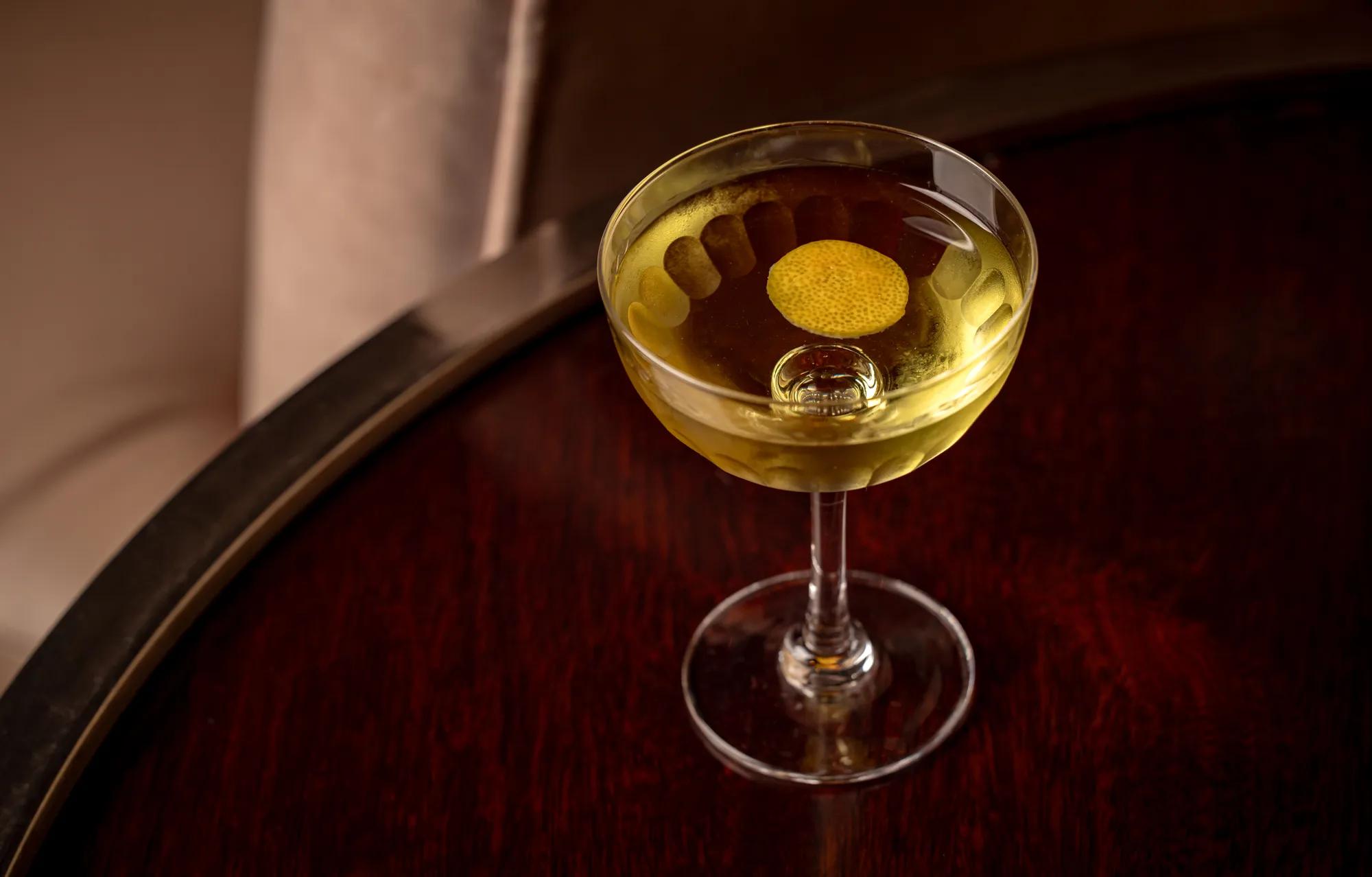 Cocktail with a lemon twist garnish served in a coupe glass on a polished dark wooden table.
