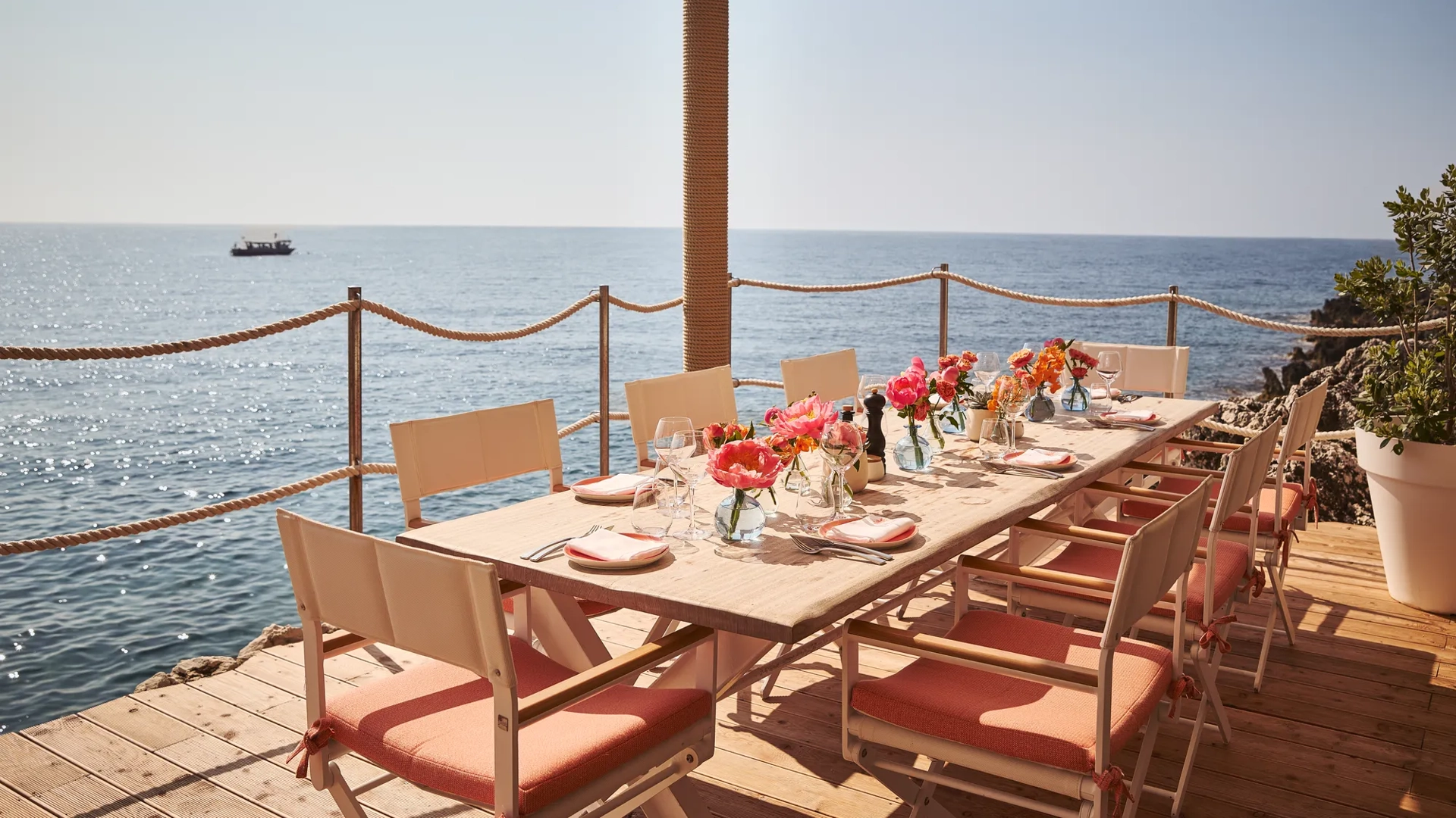 Table en bord de mer dressée avec fleurs roses, verrerie et chaises corail sous auvent face à l’océan