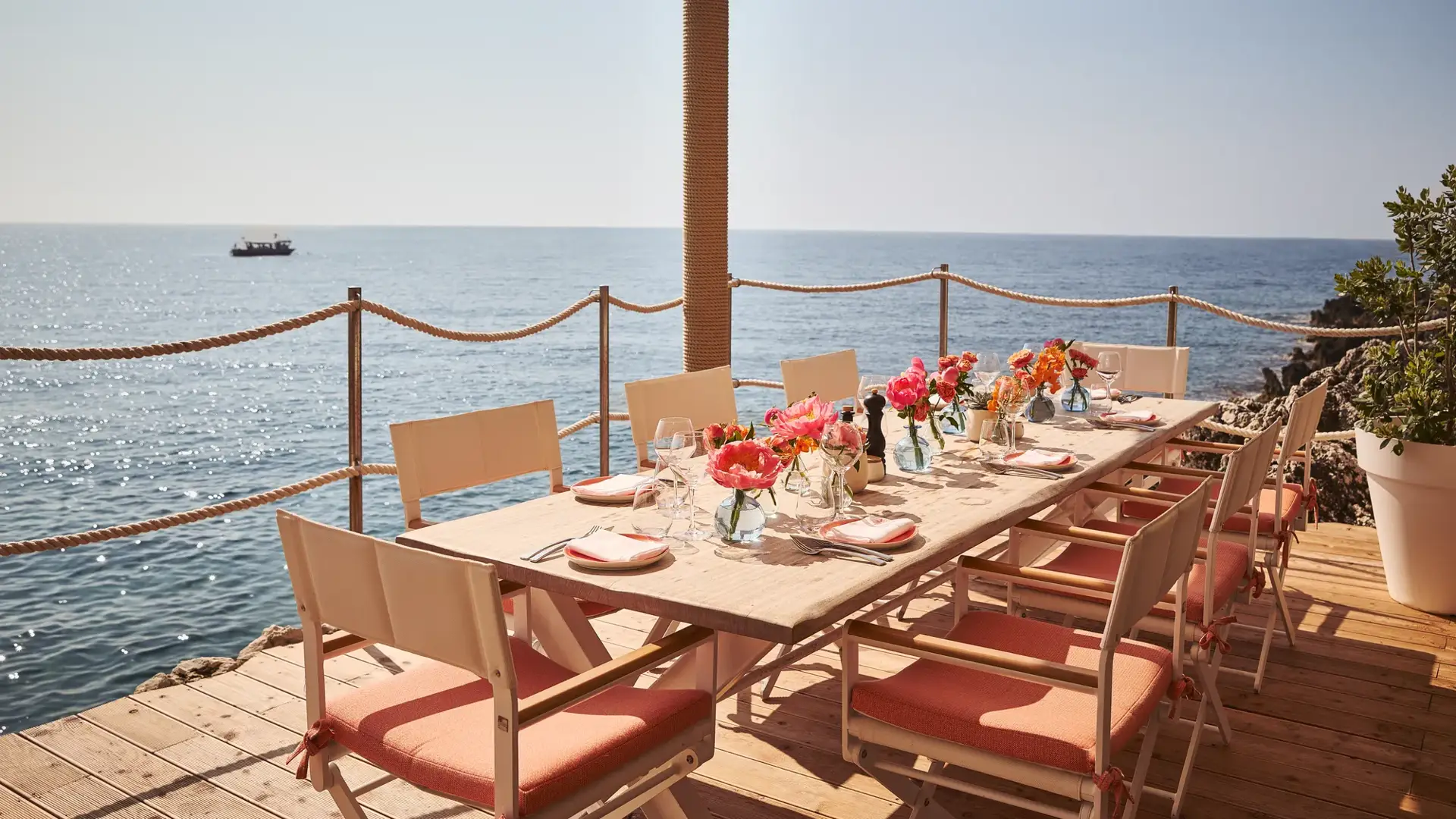 Table en bord de mer dressée avec fleurs roses, verrerie et chaises corail sous auvent face à l’océan