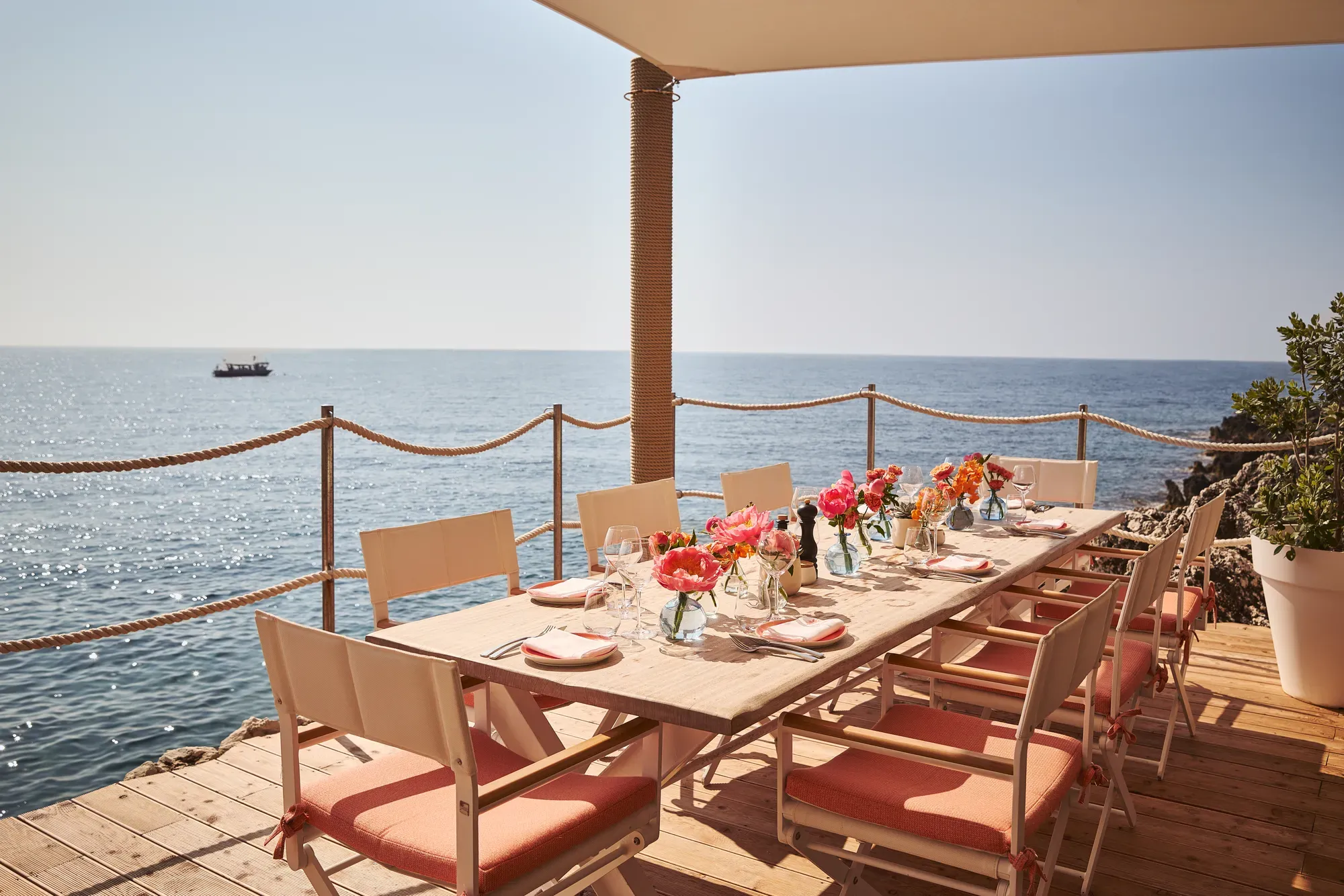 Table en bord de mer dressée avec fleurs roses, verrerie et chaises corail sous auvent face à l’océan