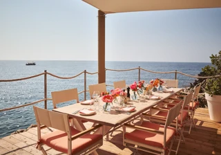 Table en bord de mer dressée avec fleurs roses, verrerie et chaises corail sous auvent face à l’océan