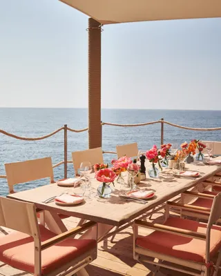 Table en bord de mer dressée avec fleurs roses, verrerie et chaises corail sous auvent face à l’océan
