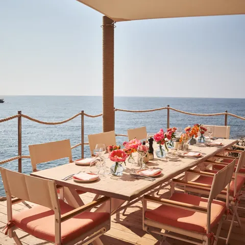 Table en bord de mer dressée avec fleurs roses, verrerie et chaises corail sous auvent face à l’océan
