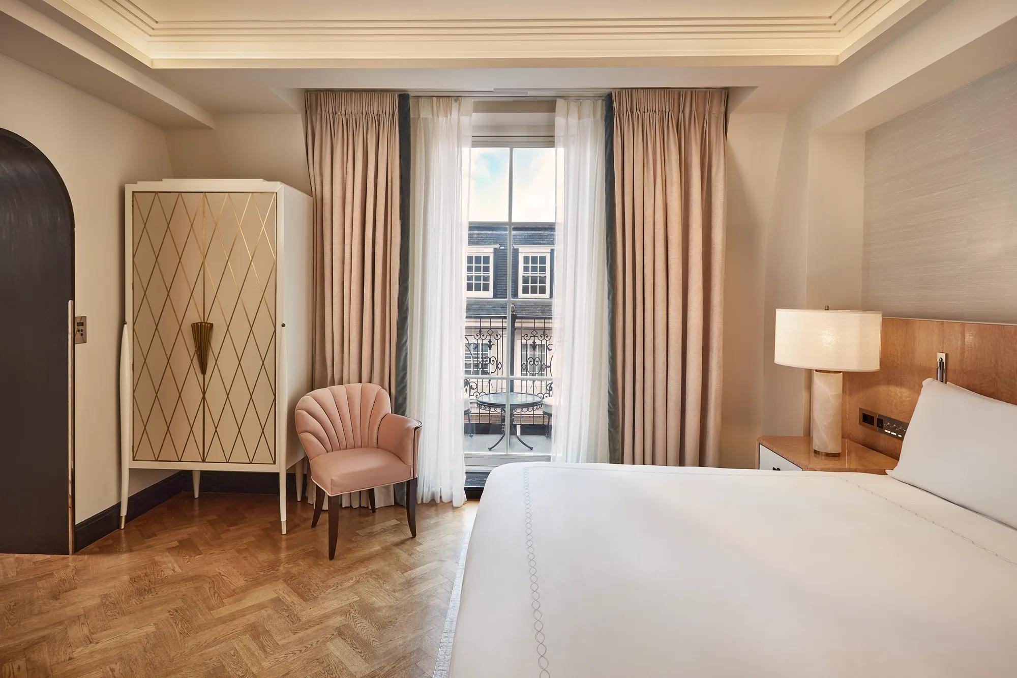 Bedroom with king bed, armchair, and wardrobe, opening onto a balcony through floor-to-ceiling curtains with city view.