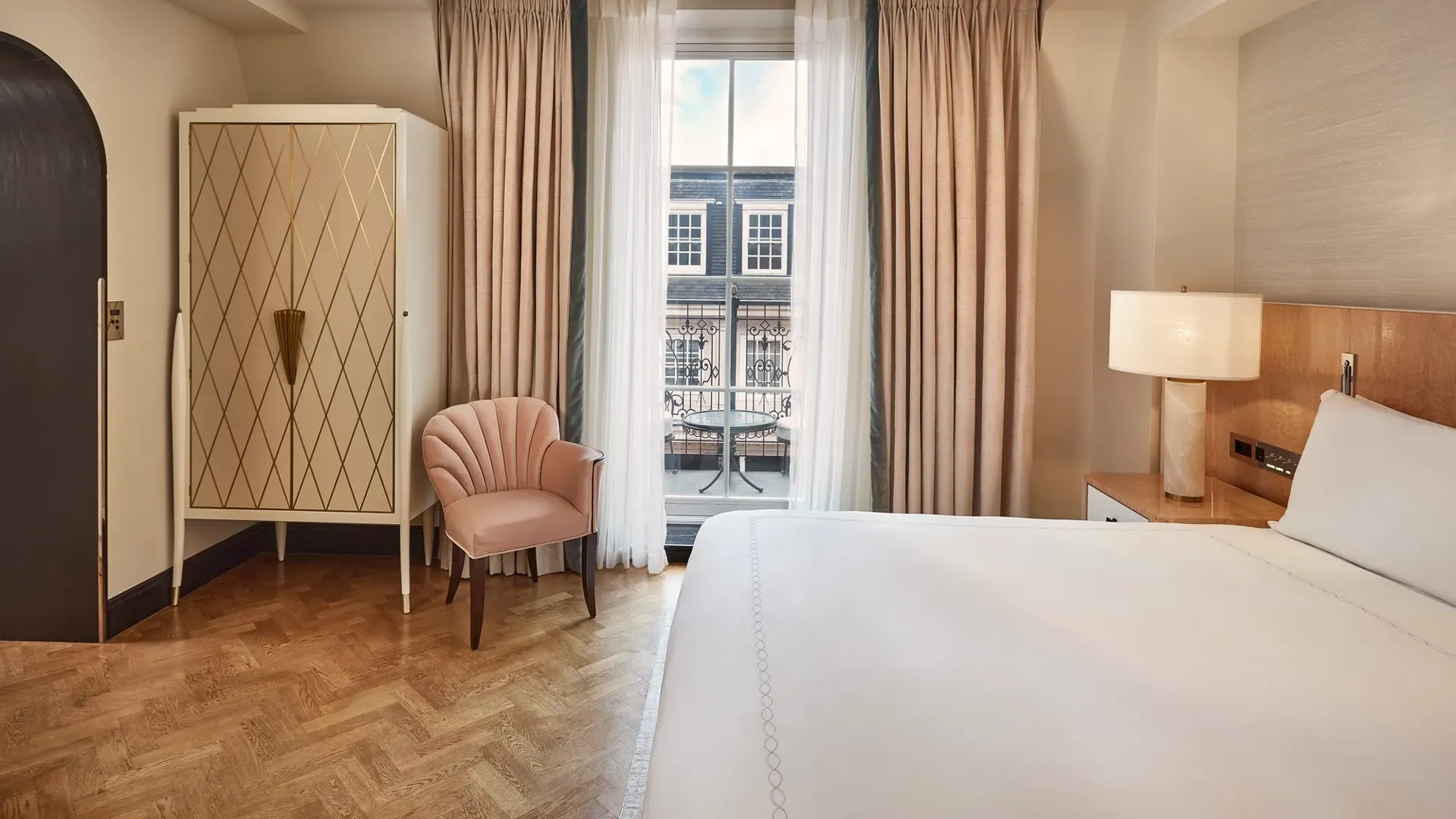Bedroom with king bed, armchair, and wardrobe, opening onto a balcony through floor-to-ceiling curtains with city view.