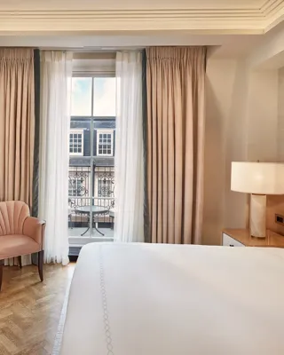 Bedroom with king bed, armchair, and wardrobe, opening onto a balcony through floor-to-ceiling curtains with city view.