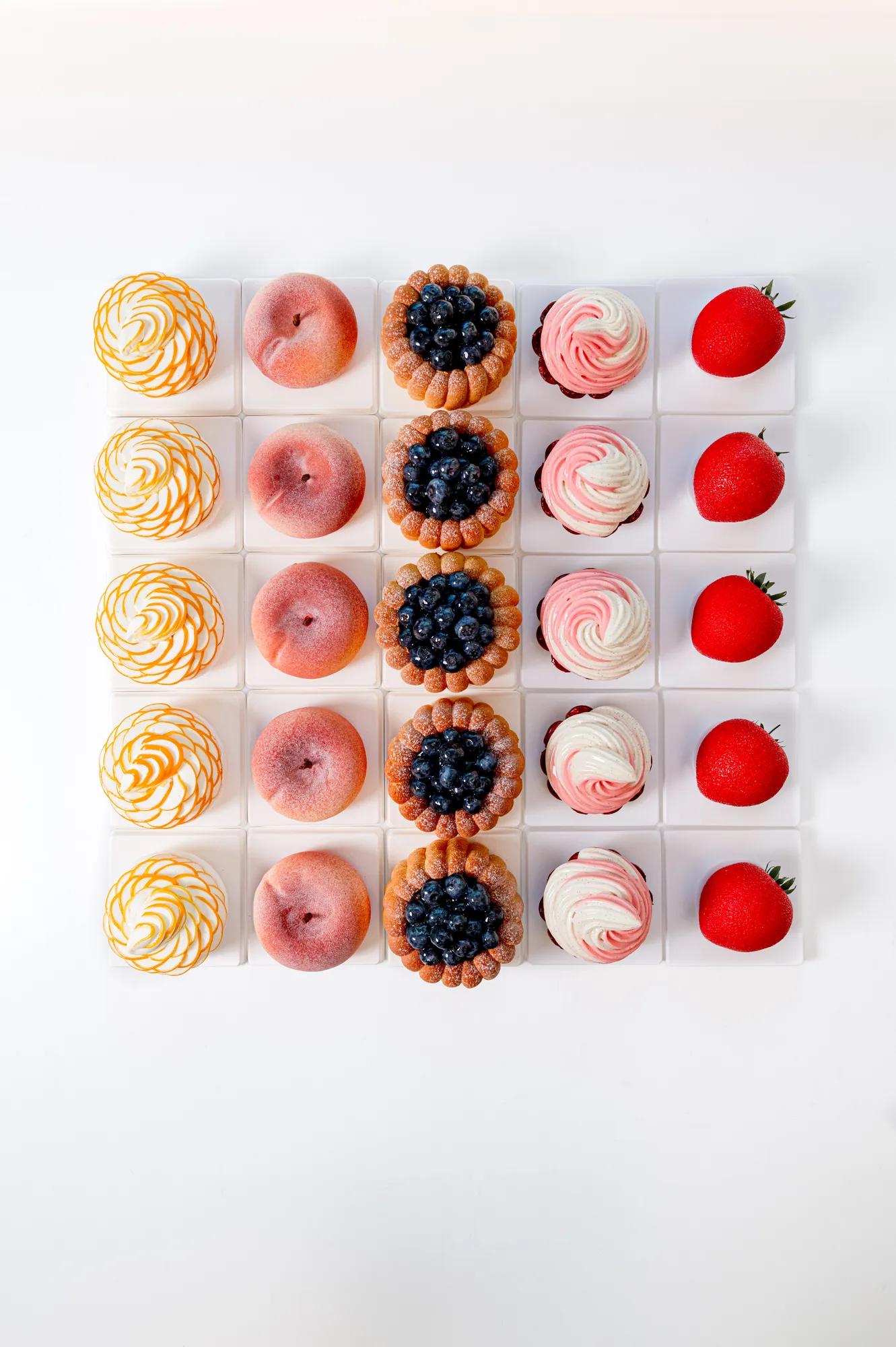 Grid arrangement of desserts featuring meringues, peach-shaped pastries, blueberry tarts and strawberries, displayed on white pedestals against a white background.