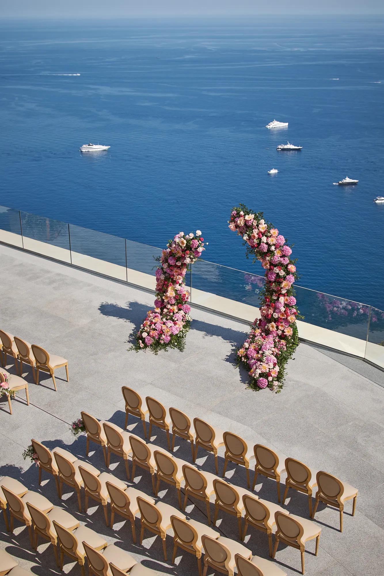 Cérémonie de mariage en bord de mer avec arches fleuries et rangées de chaises surplombant l’océan avec yachts au loin.