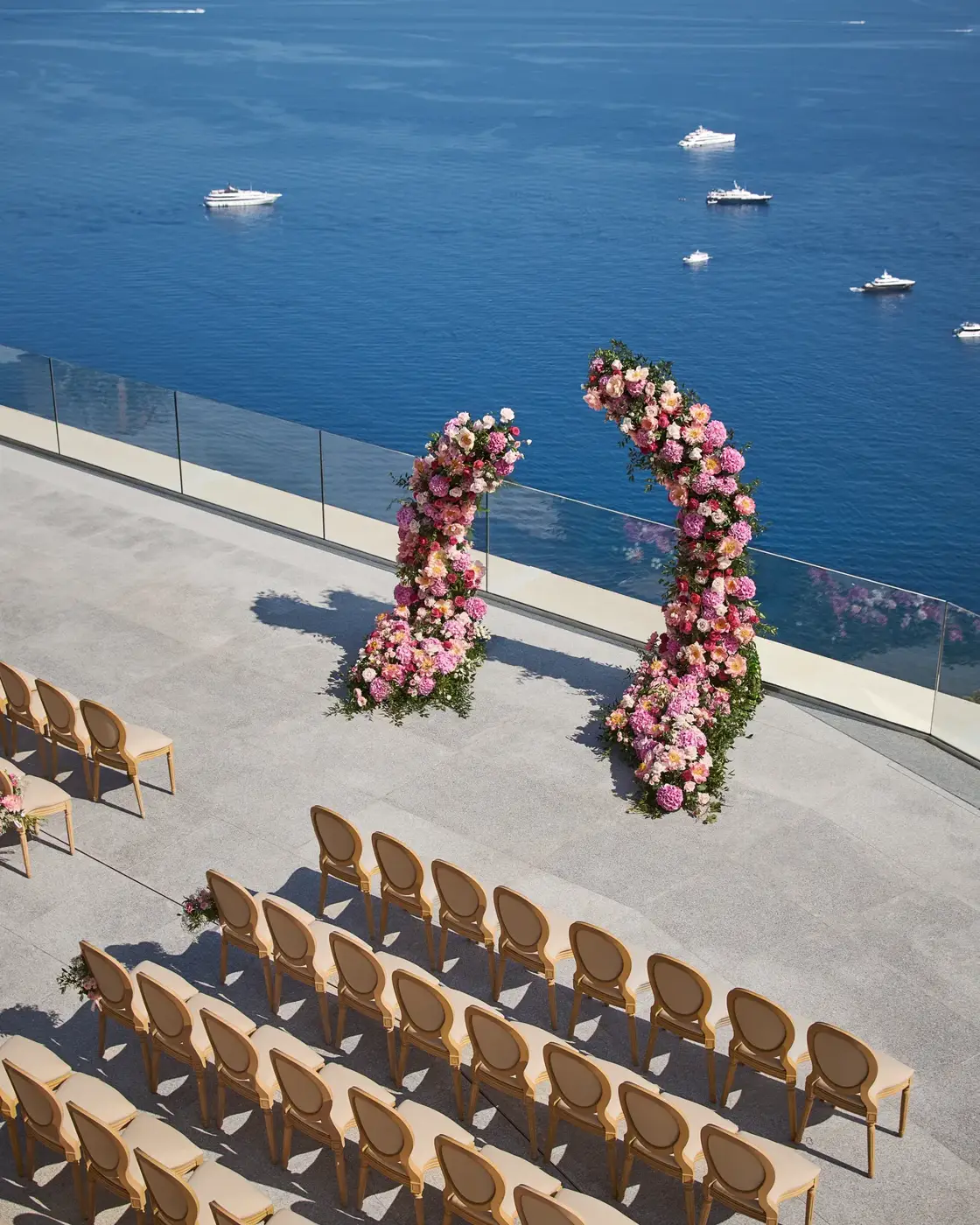 Cérémonie de mariage en bord de mer avec arches fleuries et rangées de chaises surplombant l’océan avec yachts au loin.