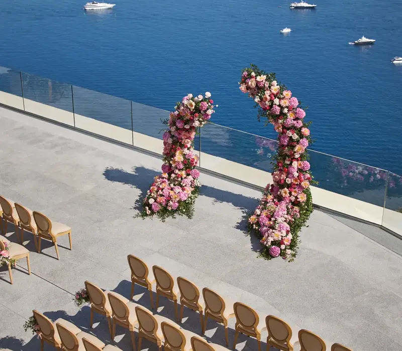 Cérémonie de mariage en bord de mer avec arches fleuries et rangées de chaises surplombant l’océan avec yachts au loin.