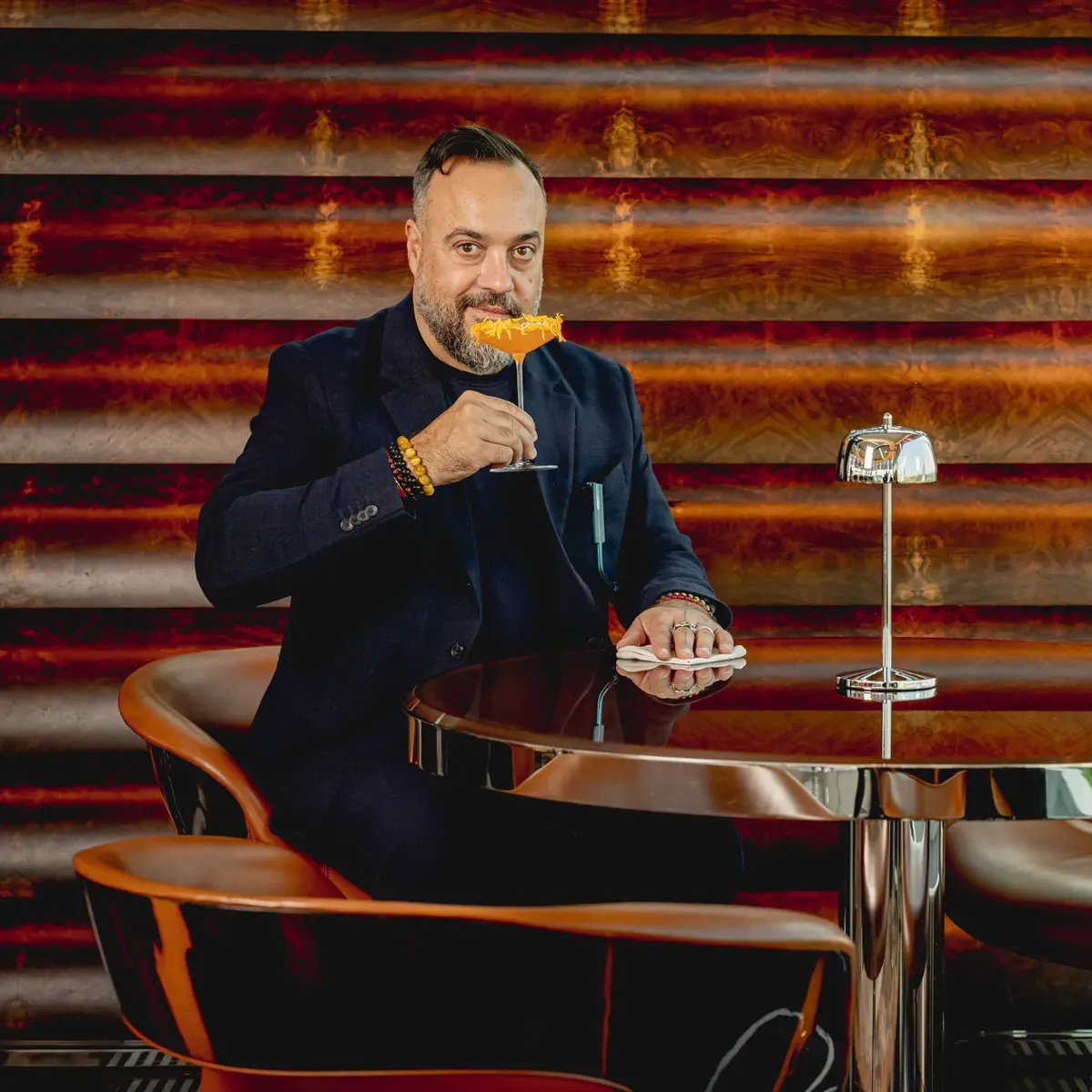 Guest seated at a bar table, holding an orange cocktail, against a patterned backdrop in a stylish lounge setting.