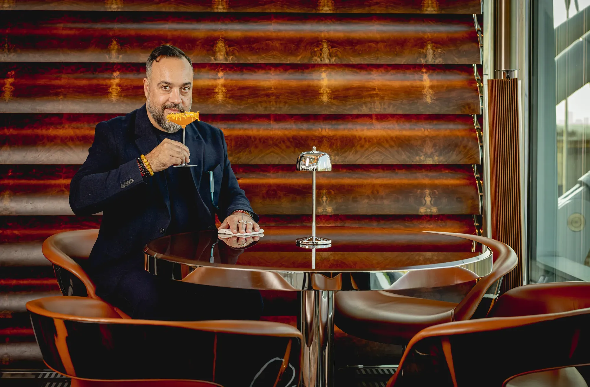 Guest seated at a bar table, holding an orange cocktail, against a patterned backdrop in a stylish lounge setting.