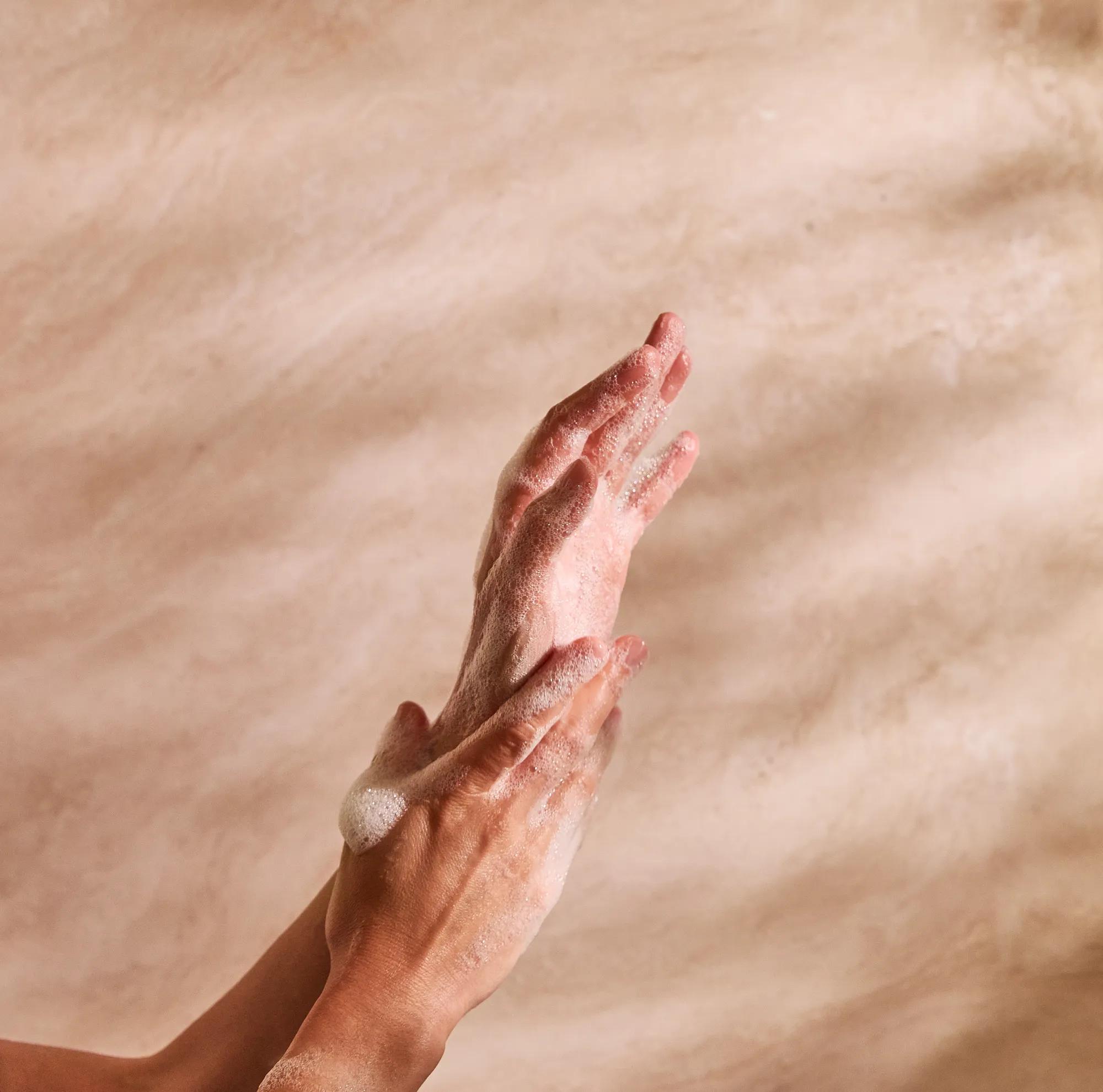 Hands lathered with soap against a warm beige stone background, highlighting a calming cleansing ritual.