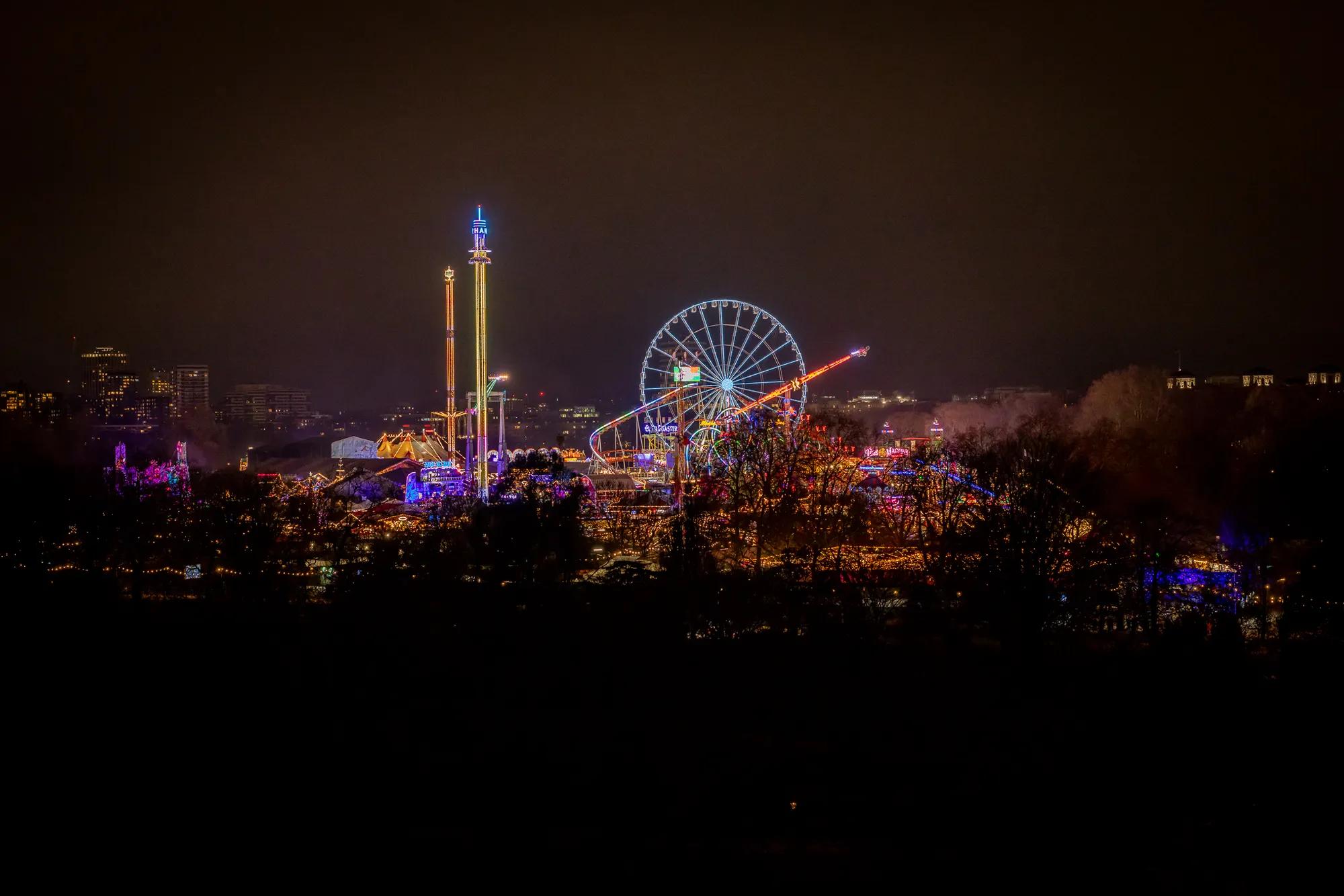 Colourfully illuminated Ferris wheel and fairground rides at night, surrounded by dark trees.