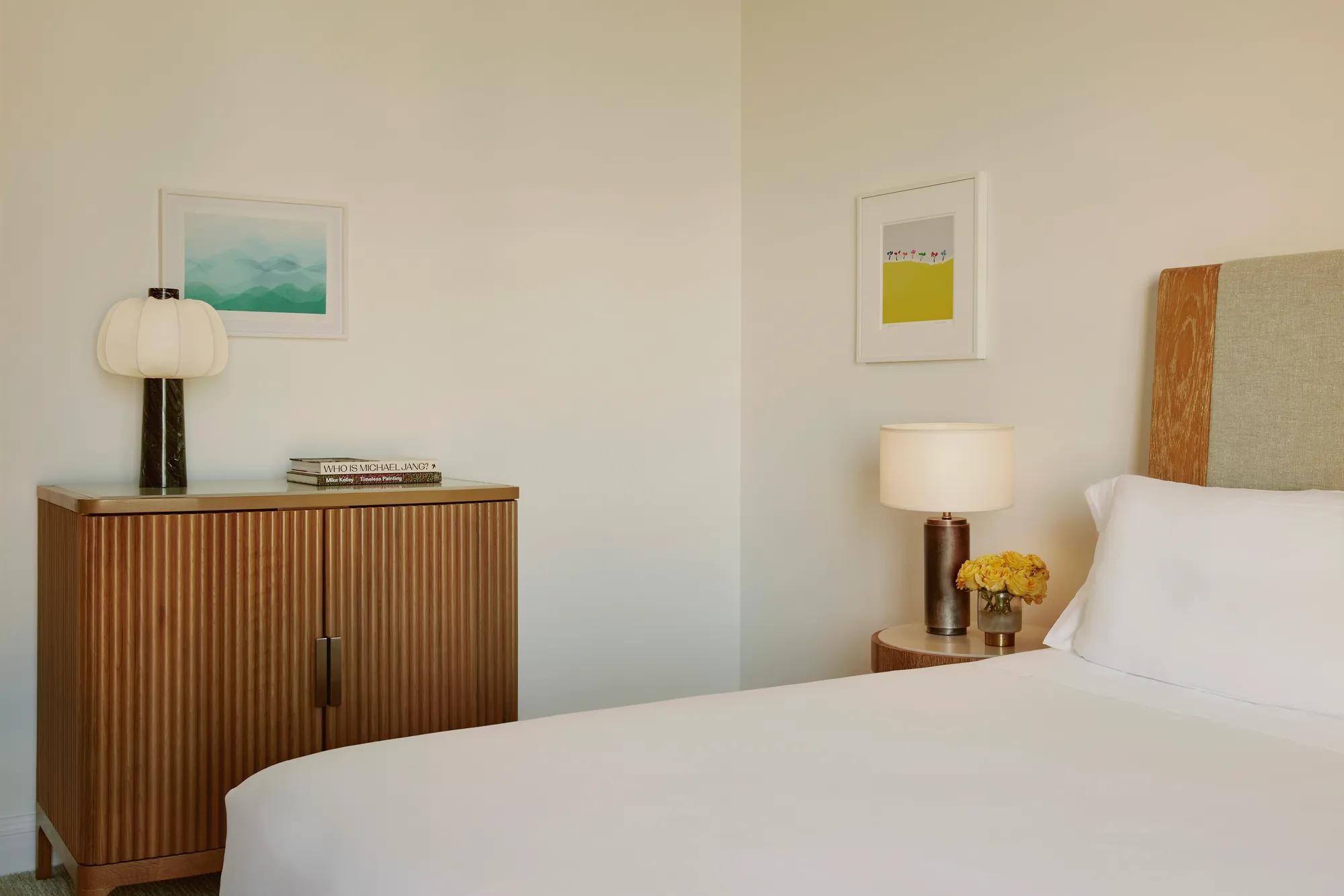 Bedroom detail showing cabinet with lamp and books, wall art, and bedside table with yellow roses.