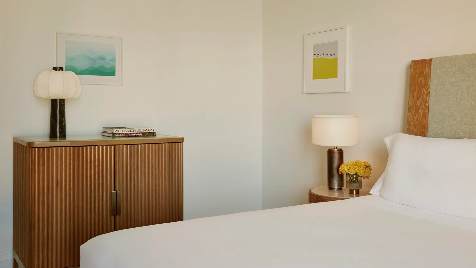 Bedroom detail showing cabinet with lamp and books, wall art, and bedside table with yellow roses.