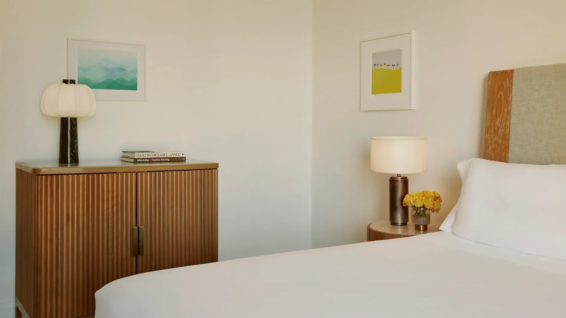 Bedroom detail showing cabinet with lamp and books, wall art, and bedside table with yellow roses.
