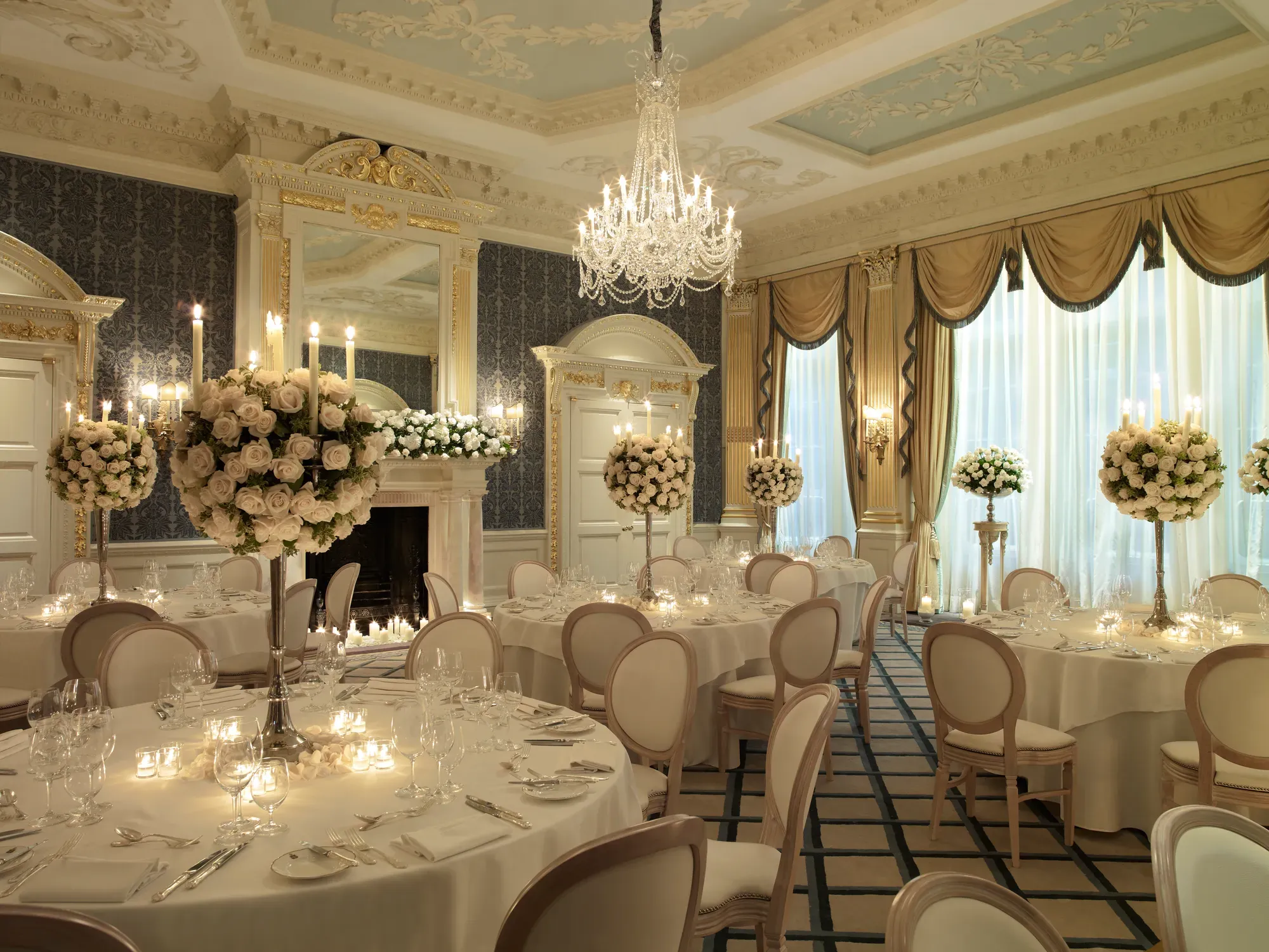 Elegant ballroom set for a formal dinner, with round tables, white linens, floral centrepieces, chandeliers, and tall windows with draped curtains.