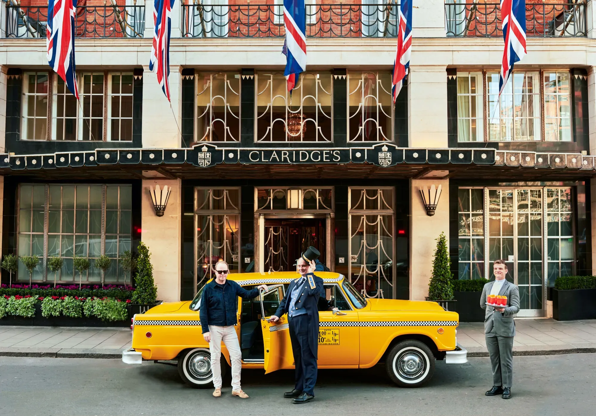 Claridge’s entrance with Union Jack flags, doorman and guest beside a yellow taxi, with staff member holding drinks nearby