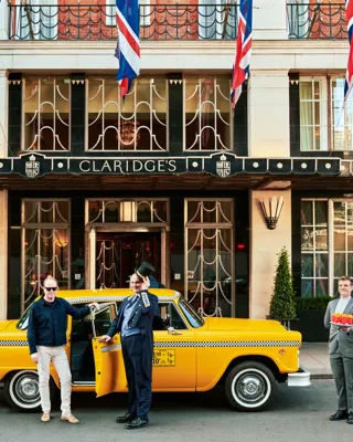 Claridge’s entrance with Union Jack flags, doorman and guest beside a yellow taxi, with staff member holding drinks nearby
