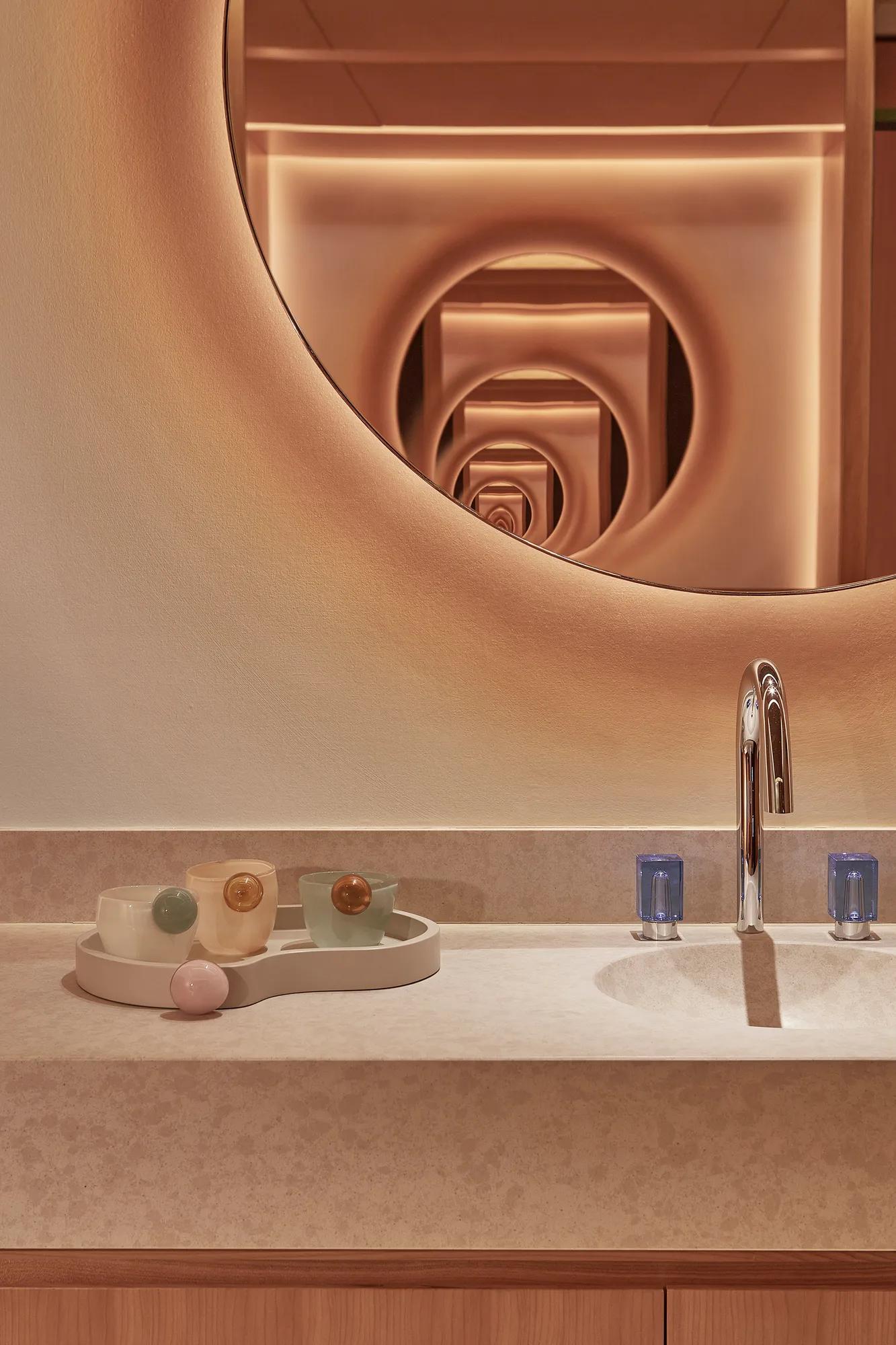 Elegant bathroom counter with soft lighting, featuring a round mirror, a sink with a polished chrome faucet, and a decorative tray with pastel-coloured cups and a small spherical object.