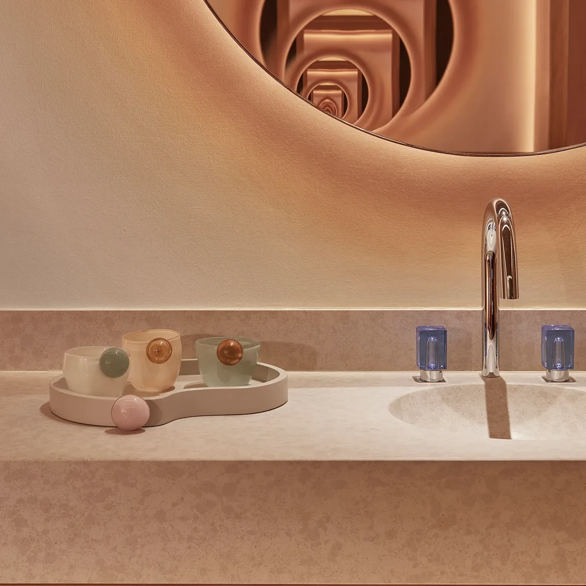 Elegant bathroom counter with soft lighting, featuring a round mirror, a sink with a polished chrome faucet, and a decorative tray with pastel-coloured cups and a small spherical object.
