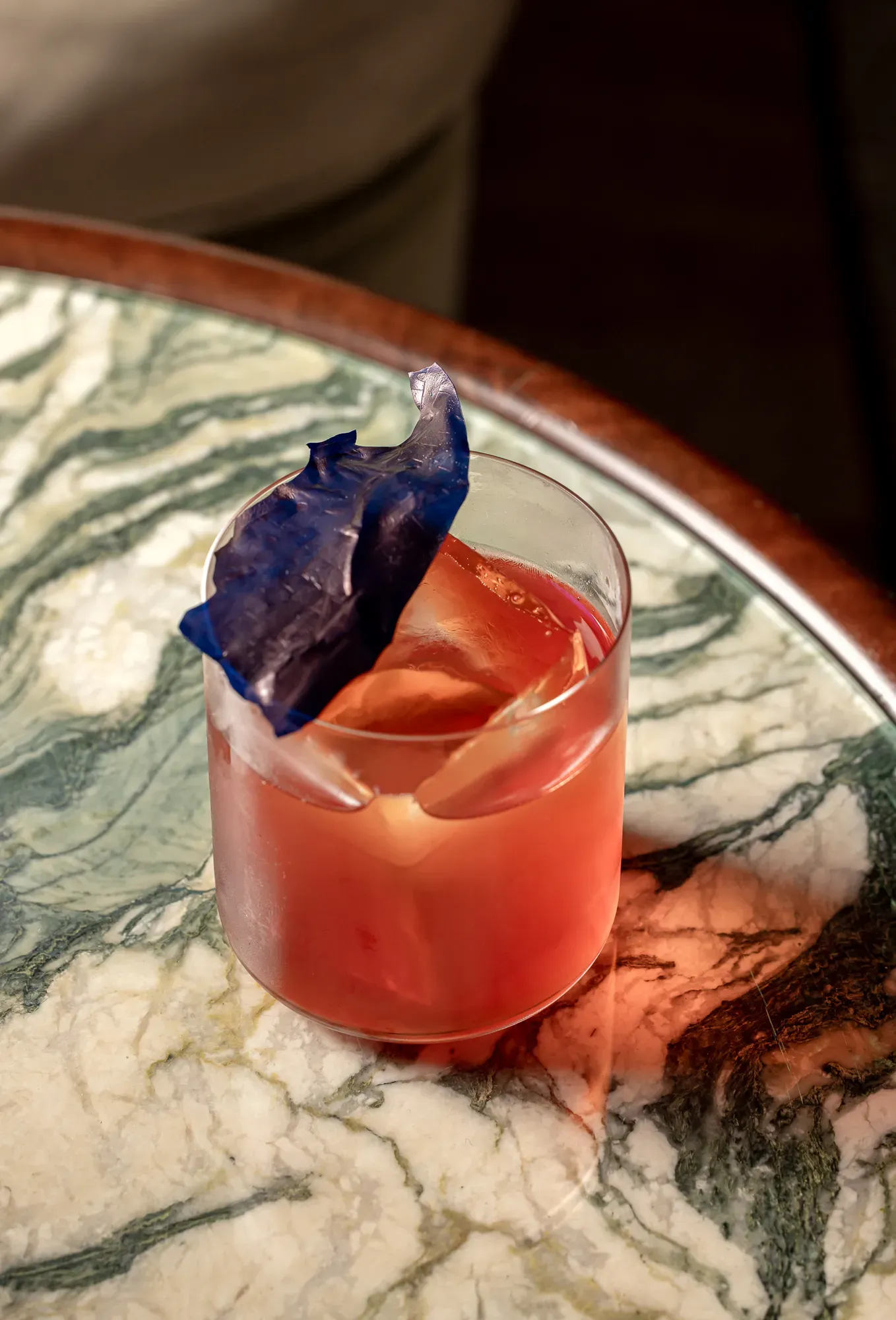 Pink cocktail in a glass with a large ice shard and a blue decorative garnish on a marbled table.