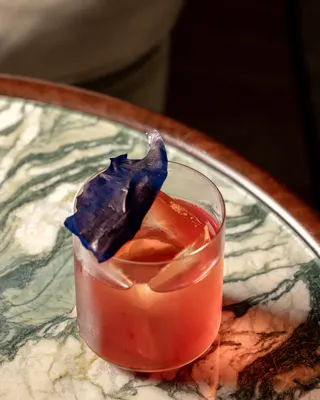 Pink cocktail in a glass with a large ice shard and a blue decorative garnish on a marbled table.