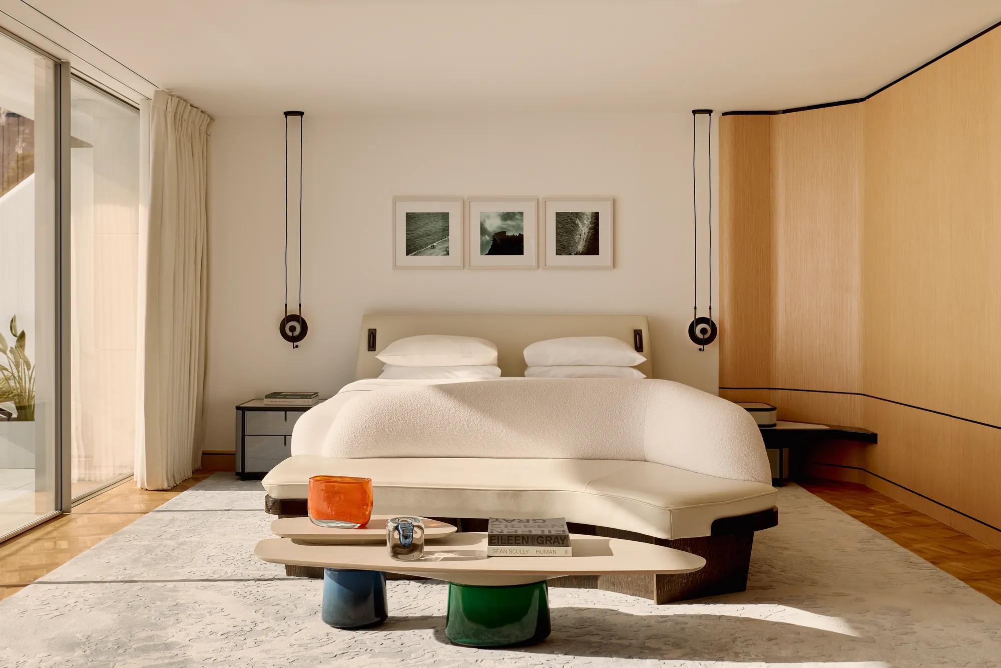 Chambre d’hôtel moderne avec grand lit, banquette incurvée et table basse, dotée de murs lambrissés et de portes vitrées s’ouvrant sur une terrasse.