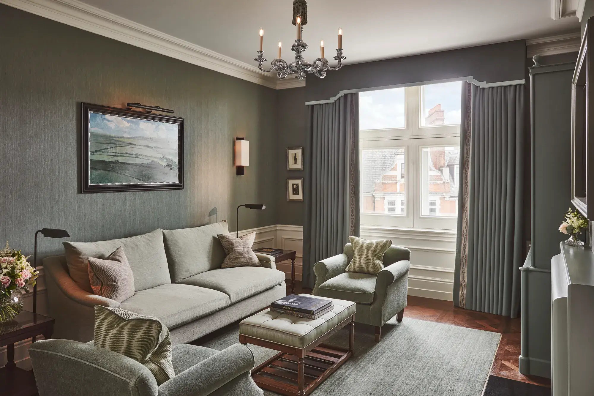 Light-filled sitting room with soft green sofa and armchairs, artwork on the wall, and a window with long drapes.
