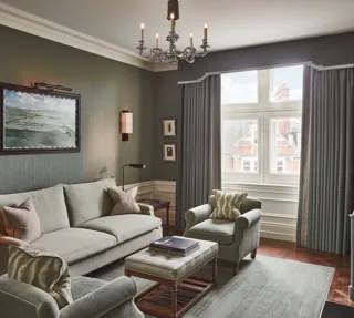 Light-filled sitting room with soft green sofa and armchairs, artwork on the wall, and a window with long drapes.