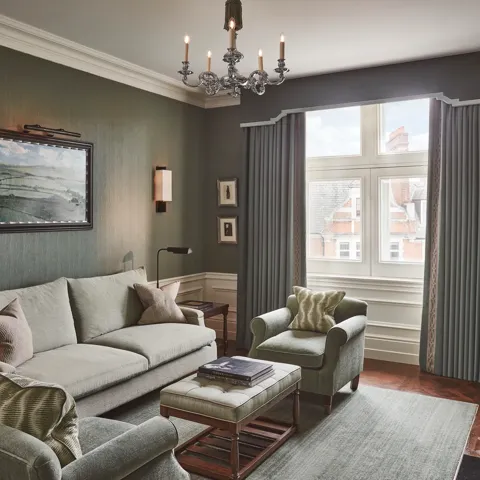 Light-filled sitting room with soft green sofa and armchairs, artwork on the wall, and a window with long drapes.