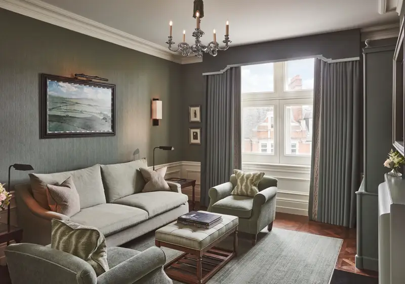 Light-filled sitting room with soft green sofa and armchairs, artwork on the wall, and a window with long drapes.