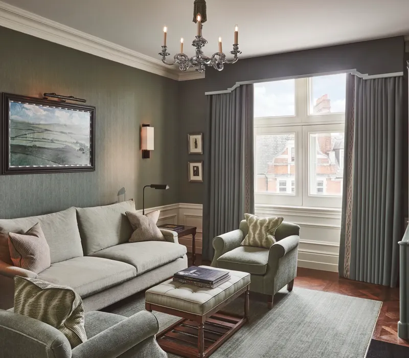 Light-filled sitting room with soft green sofa and armchairs, artwork on the wall, and a window with long drapes.