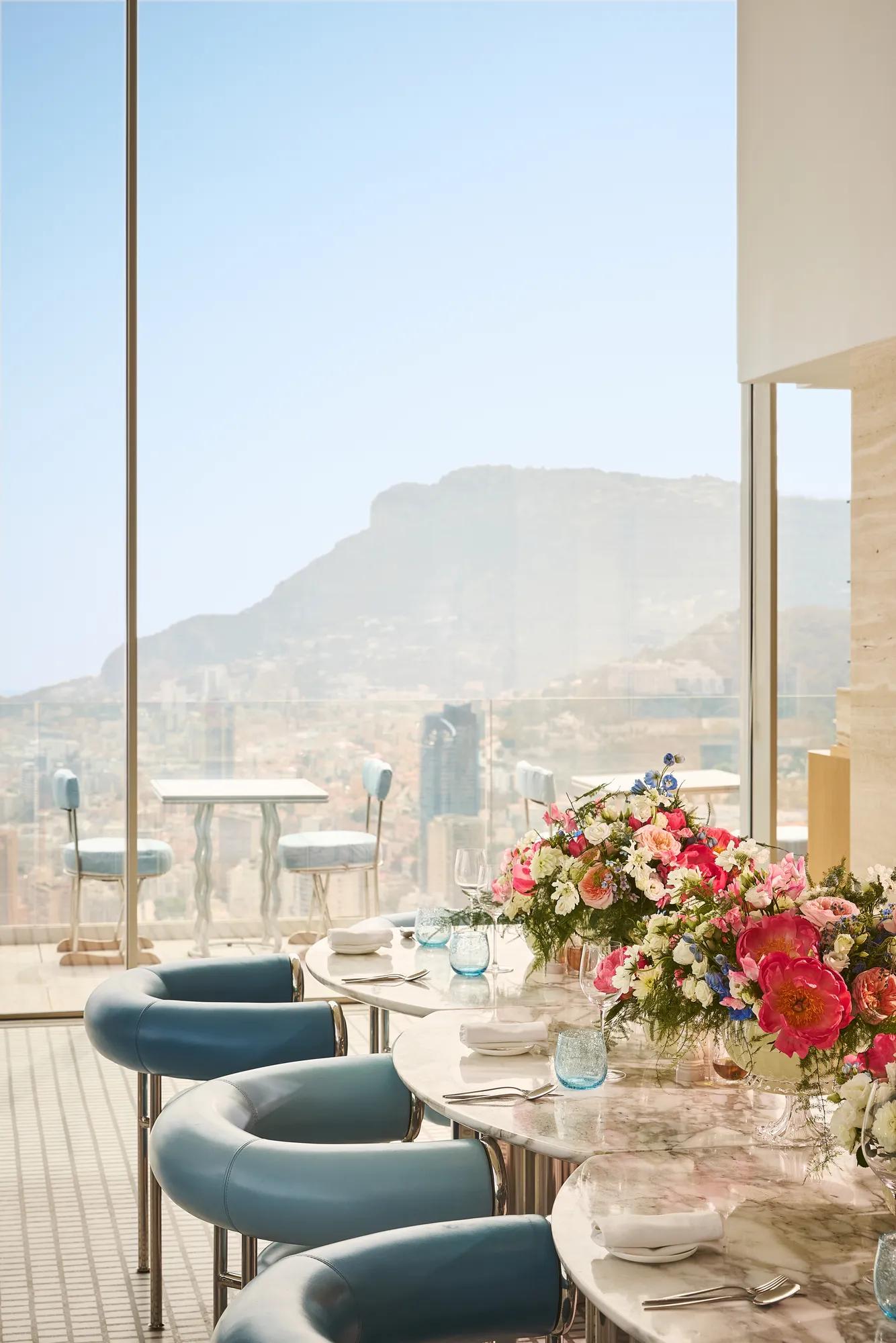 Elegant indoor dining table set with white plates, blue glassware, and a vibrant floral arrangement, beside floor-to-ceiling windows overlooking the mountains and city below.