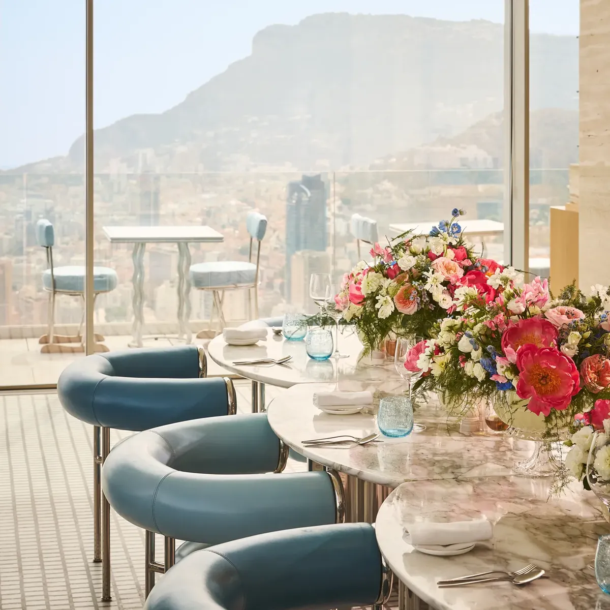 Table de salle à manger élégante dressée avec des assiettes blanches, des verres bleus et une composition florale colorée, à côté de baies vitrées offrant une vue sur la montagne et la ville en contrebas.