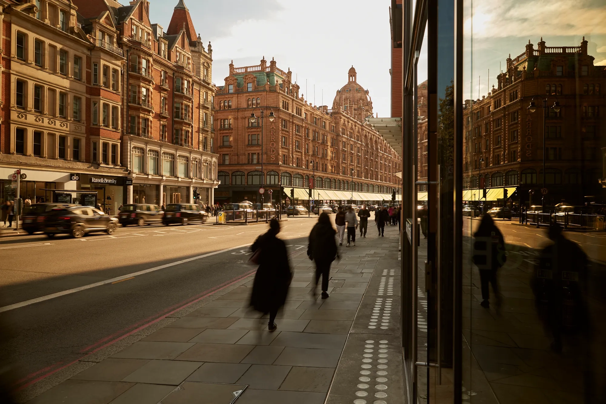 Busy London street at sunset with people walking past Harrods and traffic moving along historic buildings.