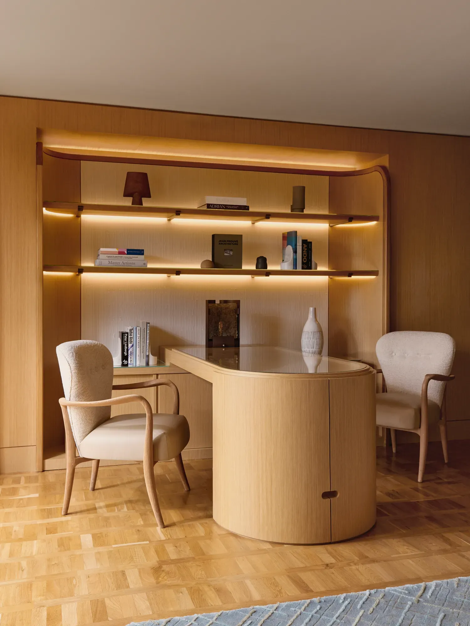 Contemporary office space with curved wooden desk, two armchairs, and backlit shelving displaying books and decorative pieces.