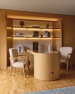 Contemporary office space with curved wooden desk, two armchairs, and backlit shelving displaying books and decorative pieces.