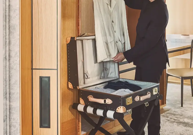Hotel staff member smiling while unpacking a beige jacket from a luxury trunk in a suite wardrobe.