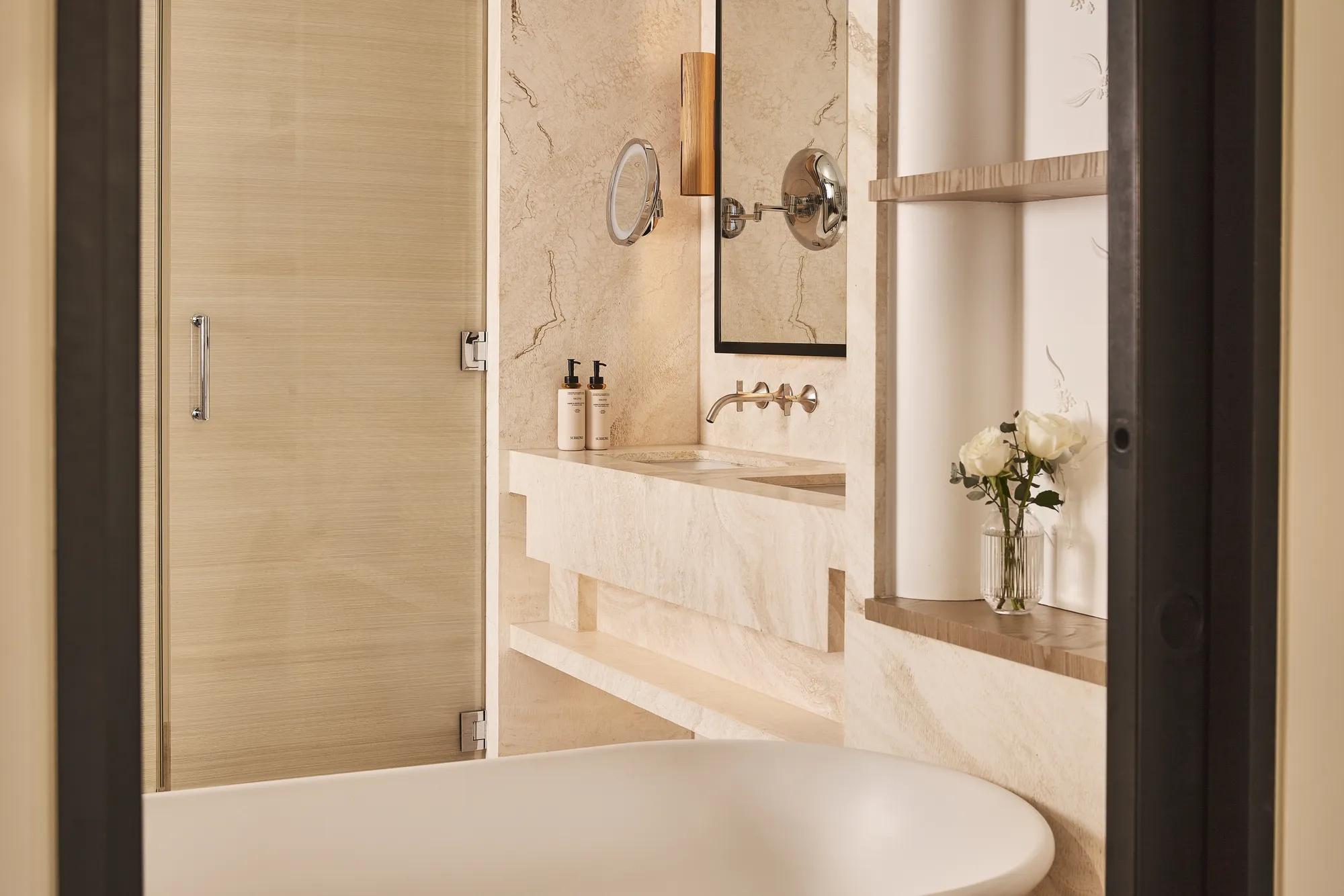 Cream stone bathroom with freestanding bathtub, wall-mounted vanity, round magnifying mirror and glass shower door.