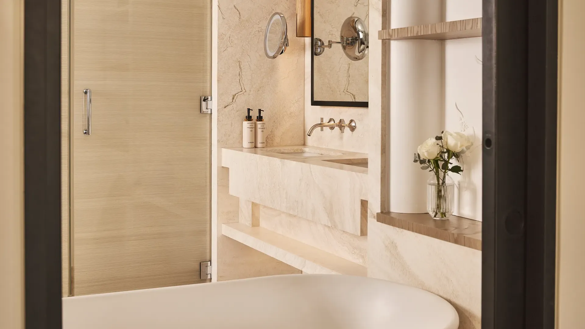 Cream stone bathroom with freestanding bathtub, wall-mounted vanity, round magnifying mirror and glass shower door.