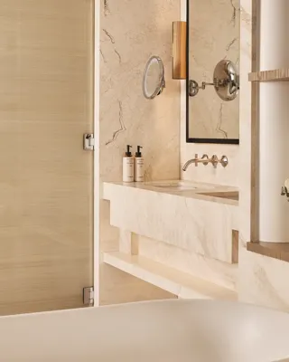 Cream stone bathroom with freestanding bathtub, wall-mounted vanity, round magnifying mirror and glass shower door.