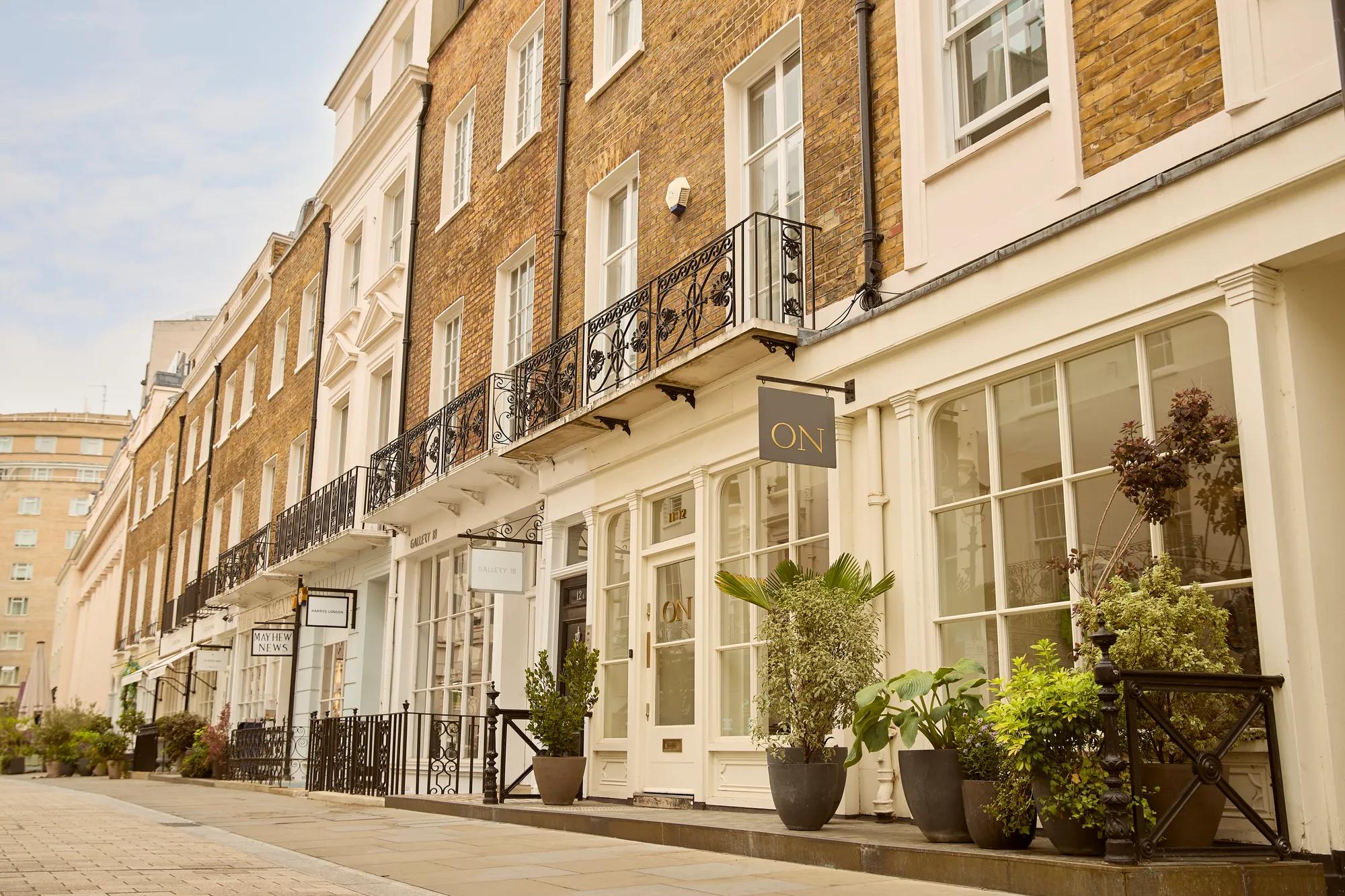 Row of elegant townhouse façades with balconies, shopfronts, and potted plants along a quiet London street.