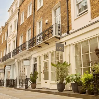 Row of elegant townhouse façades with balconies, shopfronts, and potted plants along a quiet London street.