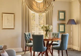Elegant dining space with a round wooden table surrounded by teal upholstered chairs, crystal chandelier above, and French doors draped with flowing curtains