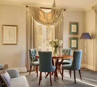 Elegant dining space with a round wooden table surrounded by teal upholstered chairs, crystal chandelier above, and French doors draped with flowing curtains